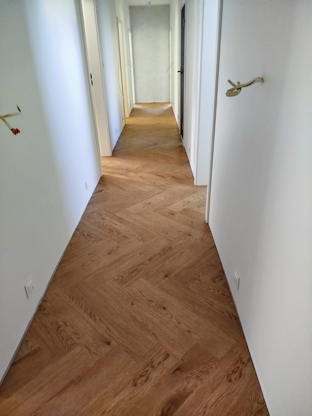 Korytarz z parkietem ułożonym w jodełkę francuską. Jasne ściany, widoczne gniazdka elektryczne. W tle otwarte drzwi do kolejnych pomieszczeń.