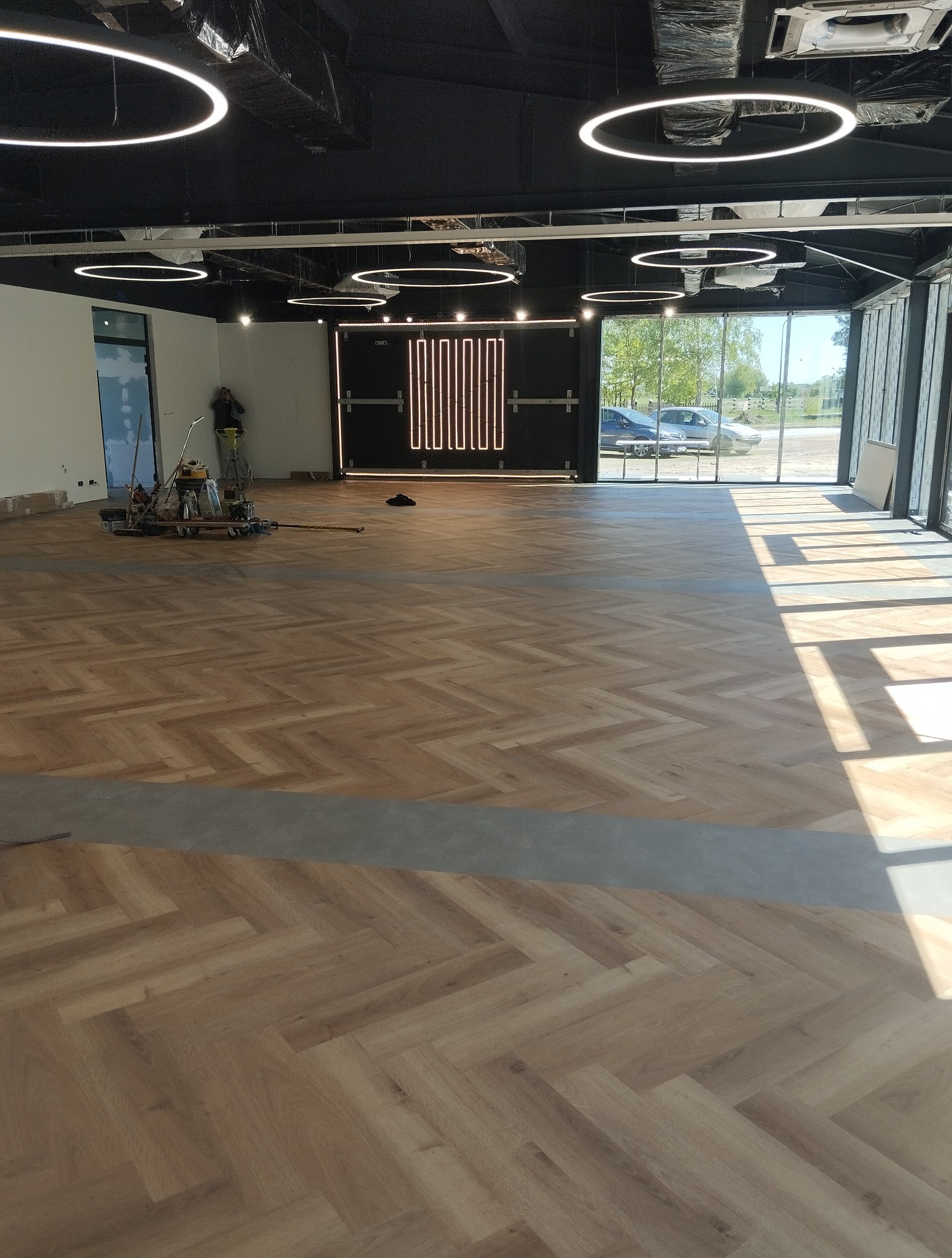 Układanie parkietu w jodełkę w przestronnym, nowoczesnym wnętrzu z oświetleniem LED. Widoczny fragment posadzki i narzędzia pracy.