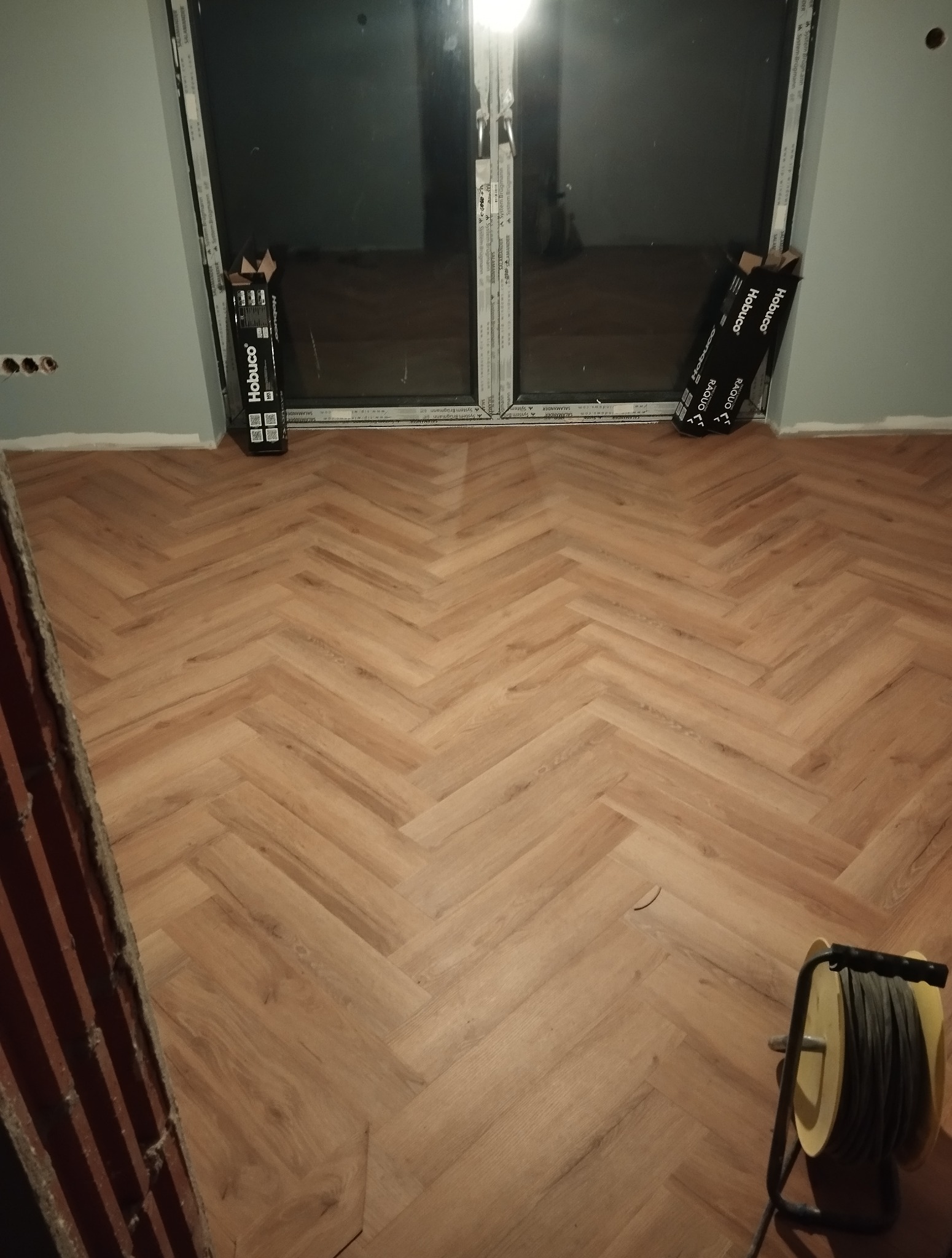 Ułożona podłoga z paneli w jodełkę, widoczne paczki paneli Hobuco przy oknie i zwinięty kabel. Fragment ściany w kolorze szarym.