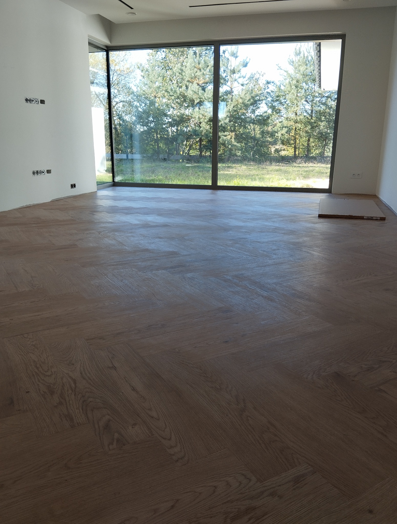 Parkiet ułożony we wzór jodełki francuskiej w jasnym, nowoczesnym wnętrzu z dużym oknem. Widoczne niedokończone fragmenty przy ścianie i karton z panelami.