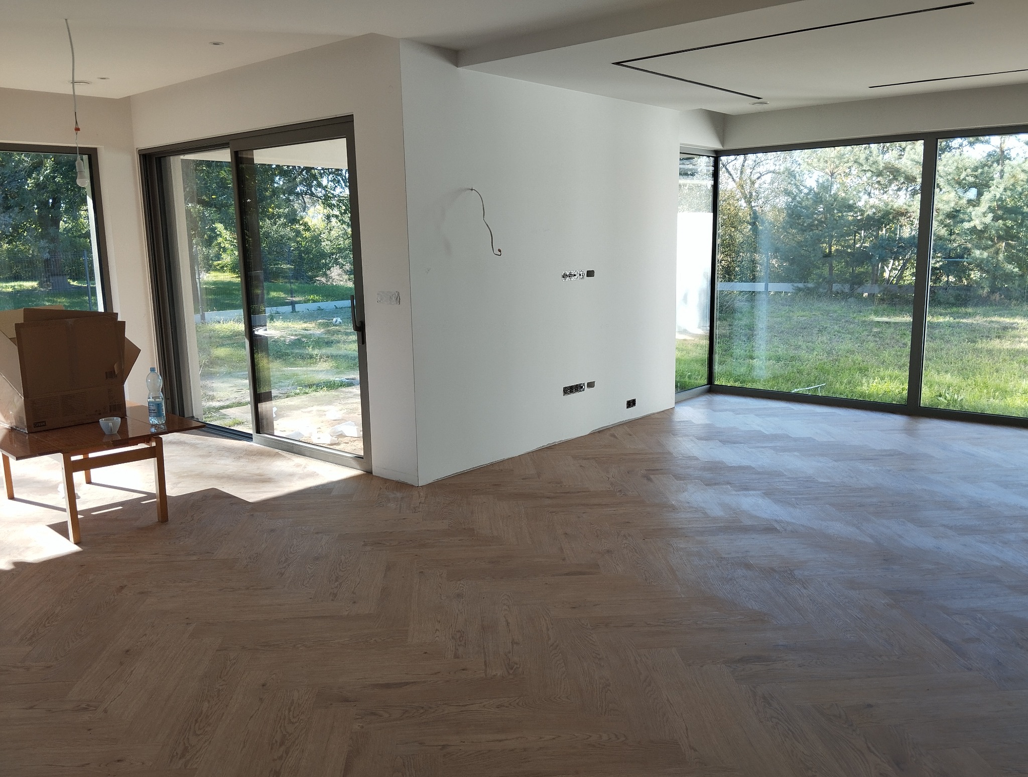 Jasne panele ułożone w jodełkę francuską w przestronnym, nowoczesnym wnętrzu z dużymi oknami. Widoczne przygotowania do wykończenia ścian.