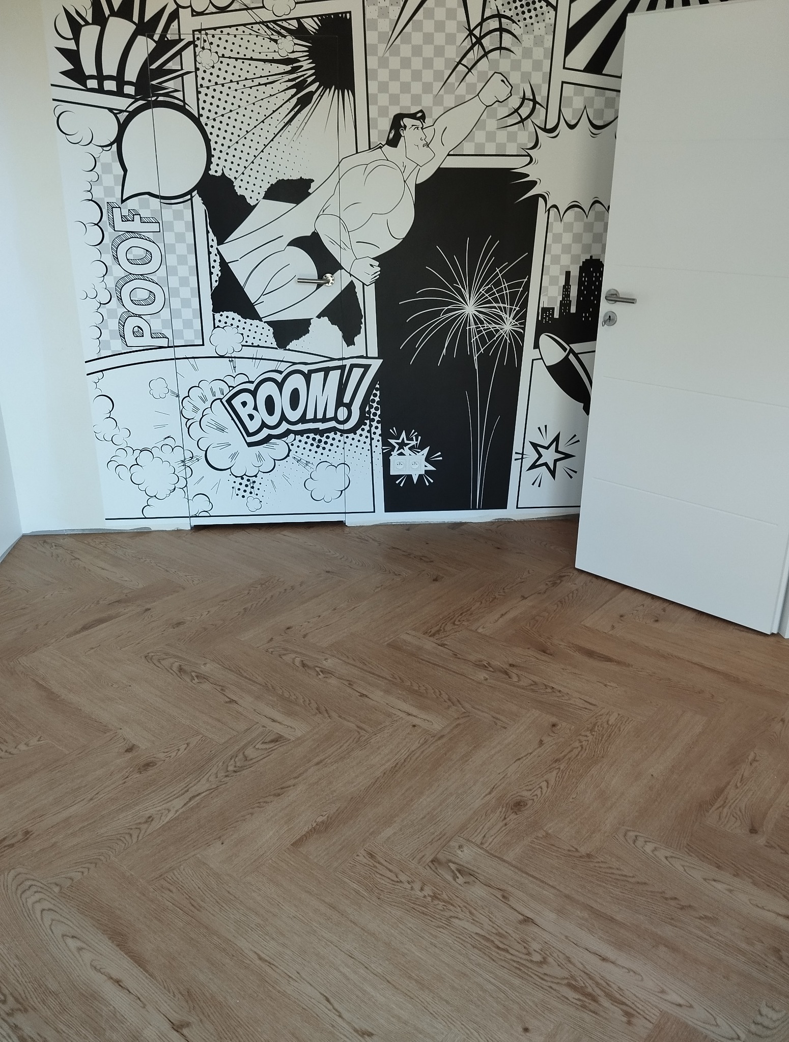 Ułożona podłoga z paneli w jodełkę francuską, jasne drewno. W tle ściana z komiksowym motywem superbohatera i białe drzwi. Widok z góry, naturalne światło.