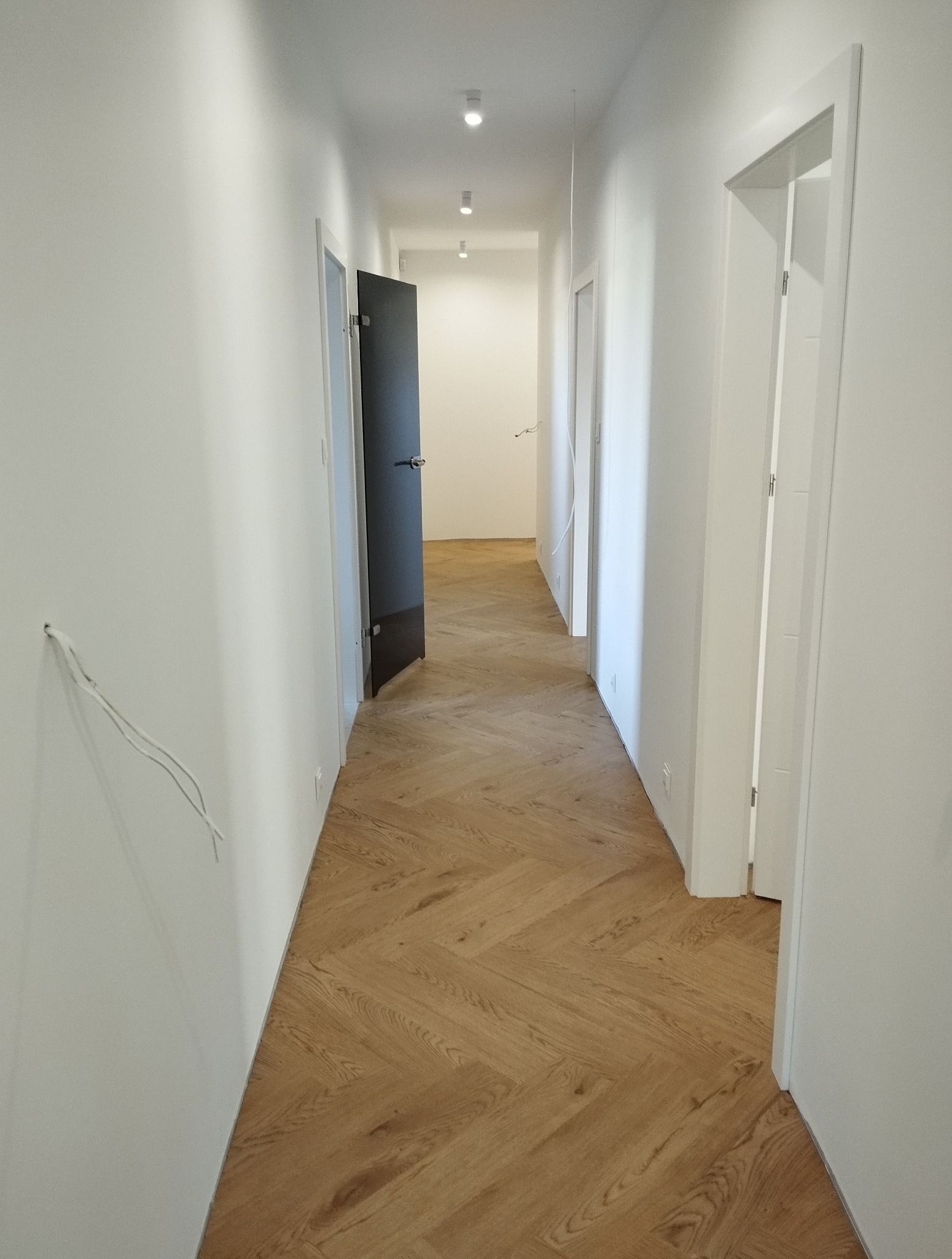 Korytarz z parkietem ułożonym w jodełkę, białe ściany i drzwi, widoczne niedokończone prace elektryczne. Jasne wnętrze z nowoczesnym oświetleniem.