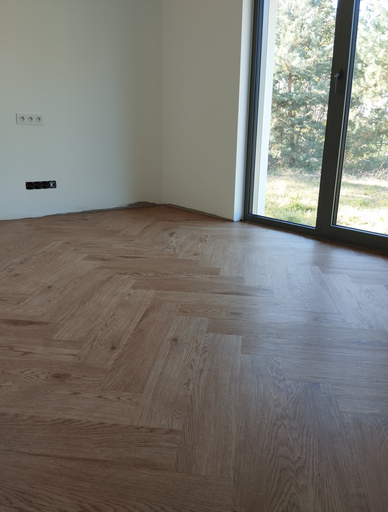 Ułożona podłoga z paneli w jodełkę w jasnym, nowoczesnym wnętrzu z dużym oknem. Widoczne gniazdka elektryczne i listwy przypodłogowe.