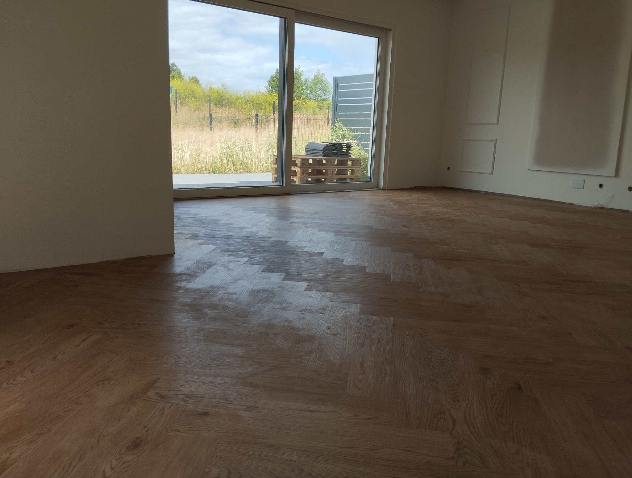 Ułożona podłoga z paneli w jodełkę w jasnym, pustym pokoju z dużym oknem przesuwnym i paletą z panelami na zewnątrz. Widok na zewnątrz z zielenią i szarym ogrodzeniem.