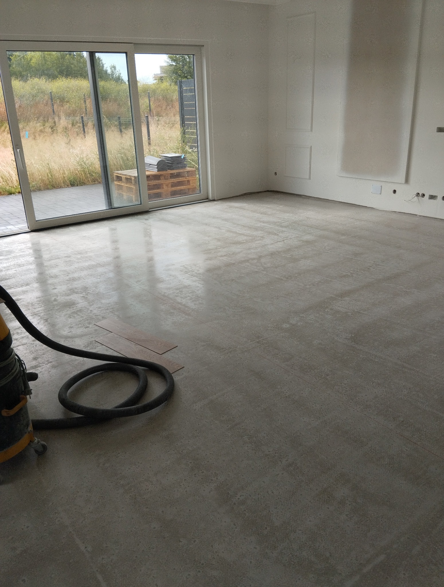 Przygotowana, szlifowana posadzka w jasnym odcieniu szarości, z widocznym odkurzaczem przemysłowym i kilkoma panelami podłogowymi, gotowa do montażu.