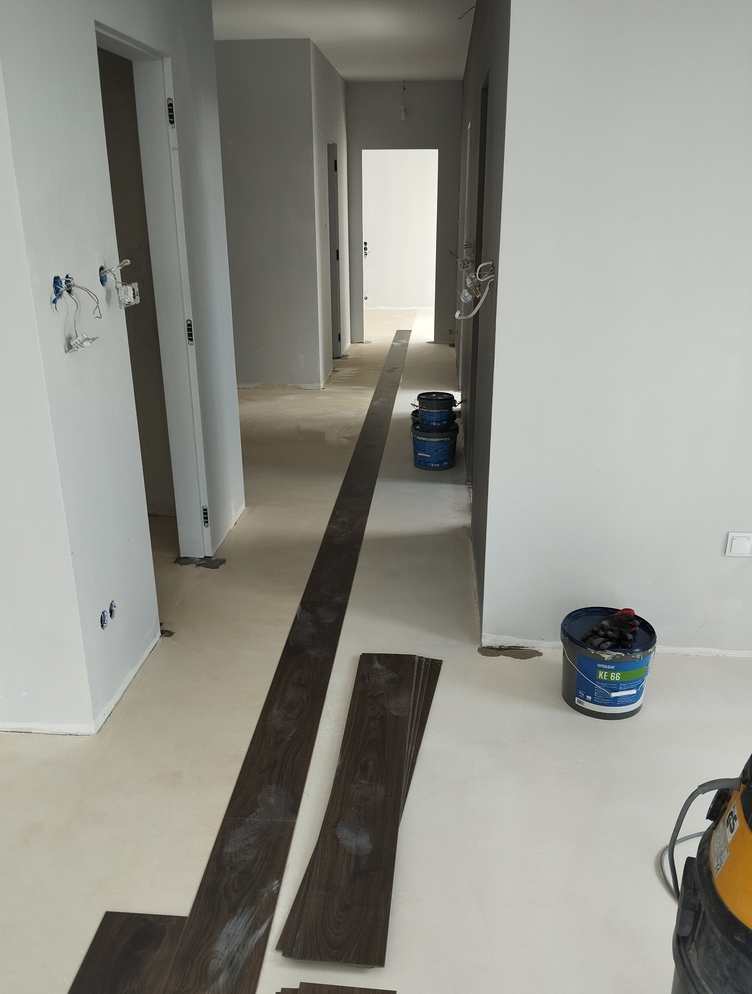 Wnętrze w trakcie układania paneli podłogowych. Widoczne panele w kolorze ciemnego drewna, puszki z klejem i odkurzacz przemysłowy. Surowe ściany i instalacja elektryczna.