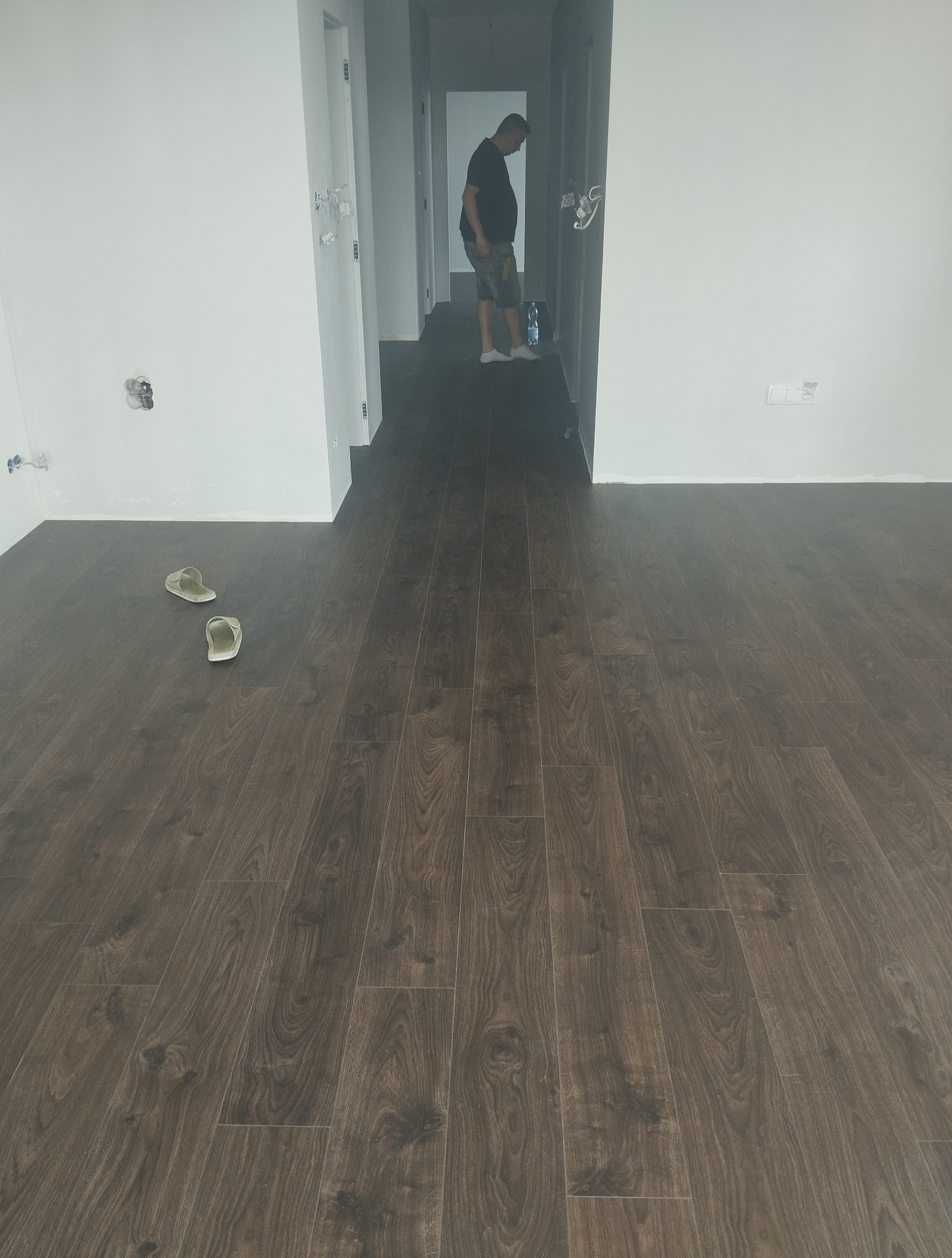 Ułożona, ciemna podłoga z paneli w nowym mieszkaniu. Widoczne białe ściany, drzwi i para klapek. W tle sylwetka mężczyzny. Układanie paneli.