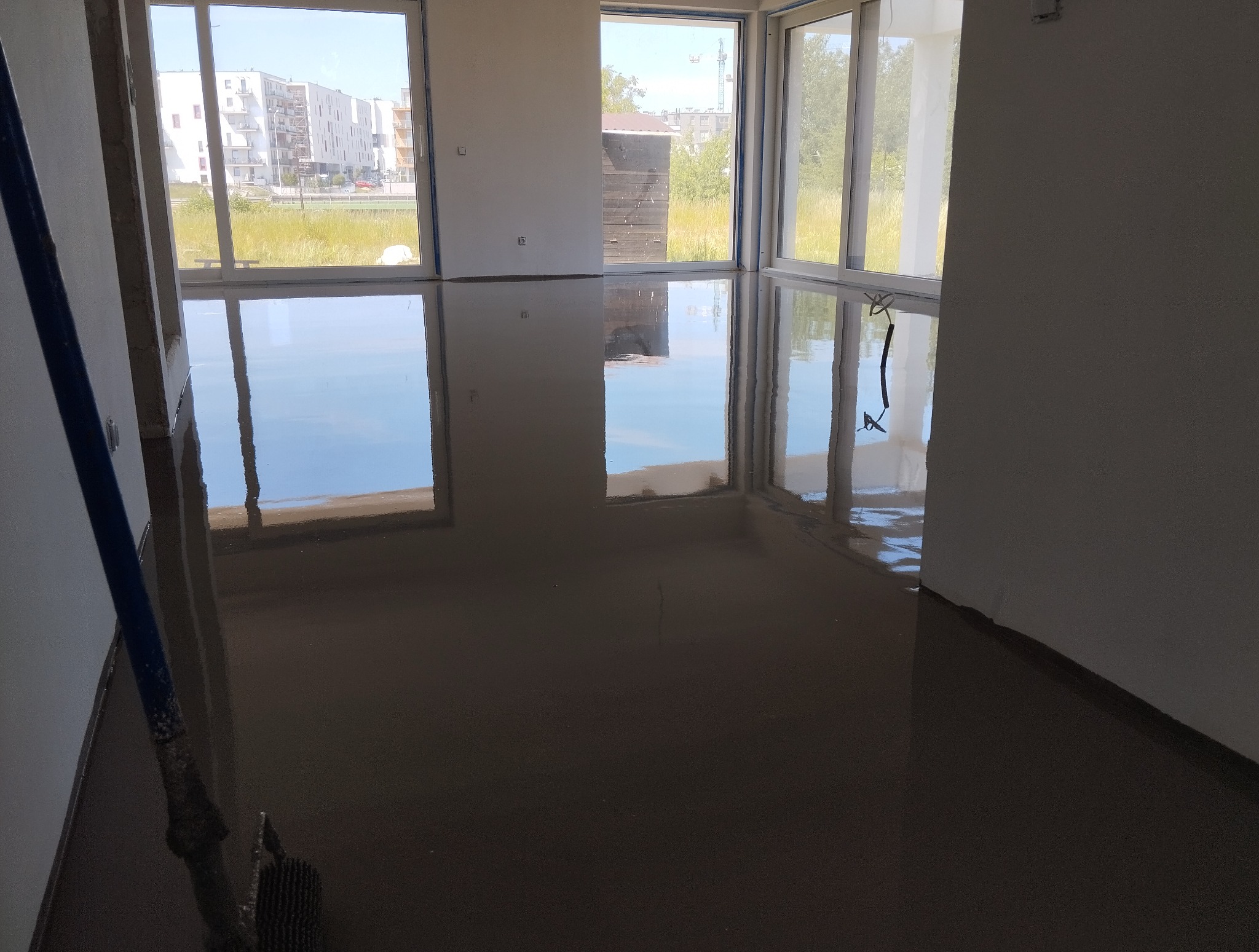 Wylewka samopoziomująca w Starej Wsi, świeżo wykonana, odbijająca okna i niebo. Widoczny fragment narzędzia do rozprowadzania masy.