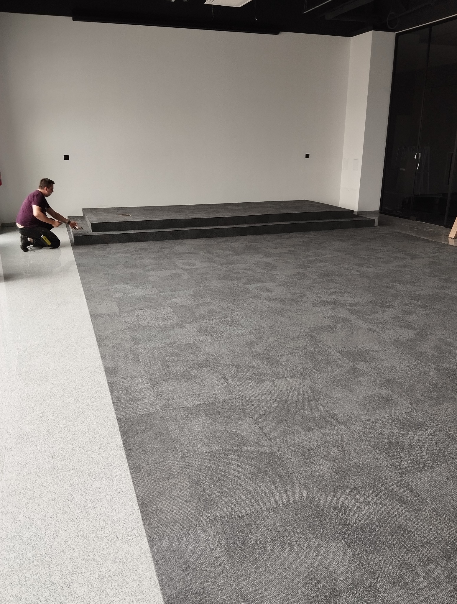 Montaż szarej wykładziny dywanowej w dużym pomieszczeniu z podestem. Pracownik docina wykładzinę przy ścianie. Część podłogi wyłożona jasną wykładziną.