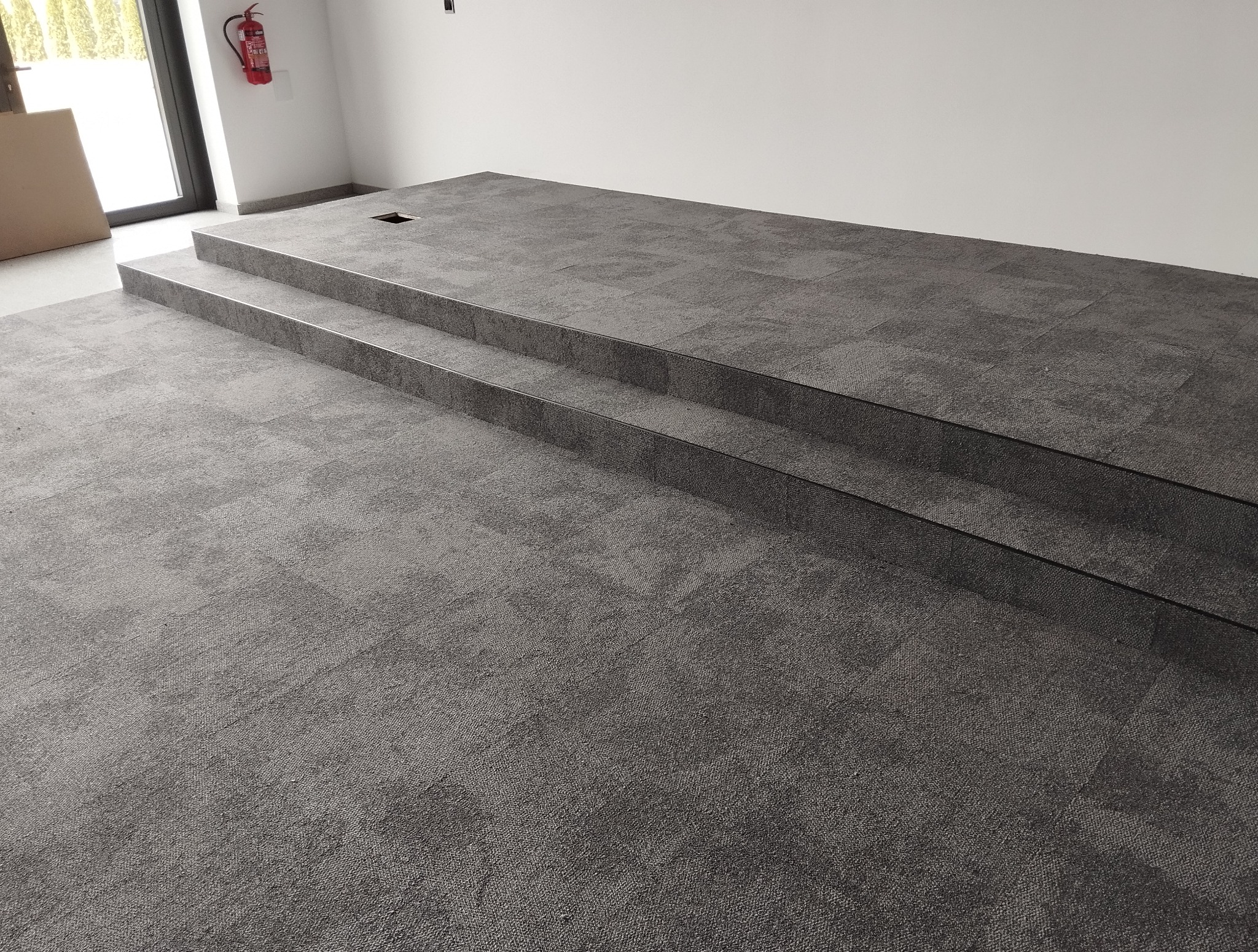 Nowoczesna, dwustopniowa podłoga wyłożona szarą wykładziną dywanową w nowym budynku. Widoczny fragment ściany i okna. Wykończenie listwami.