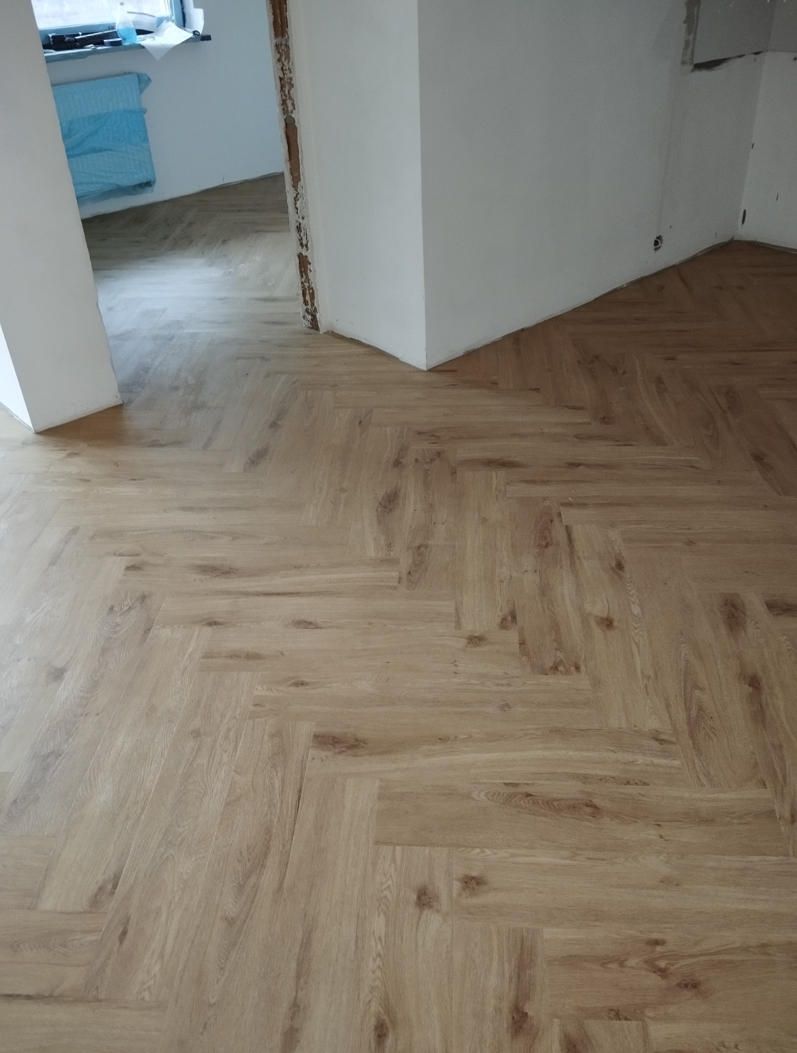 Ułożona podłoga z paneli w jodełkę, jasne drewno, widok z góry, fragment ściany z niedokończonym wykończeniem, surowe wnętrze w trakcie remontu.