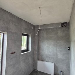 PROWORK - Szara łazienka po remoncie z płytkami na ścianach i podłodze, widoczne okno i grzejnik. Na suficie wystający kabel. Ujęcie z perspektywy narożnika.