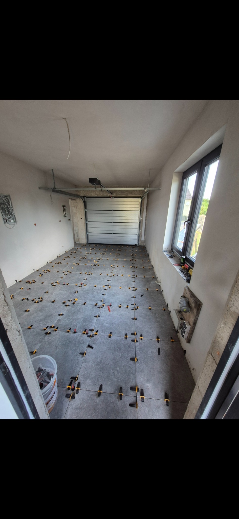 Szare płytki podłogowe w garażu, z żółtymi krzyżykami dystansowymi, okno z widokiem na zewnątrz, brama garażowa w tle, surowy stan ścian.