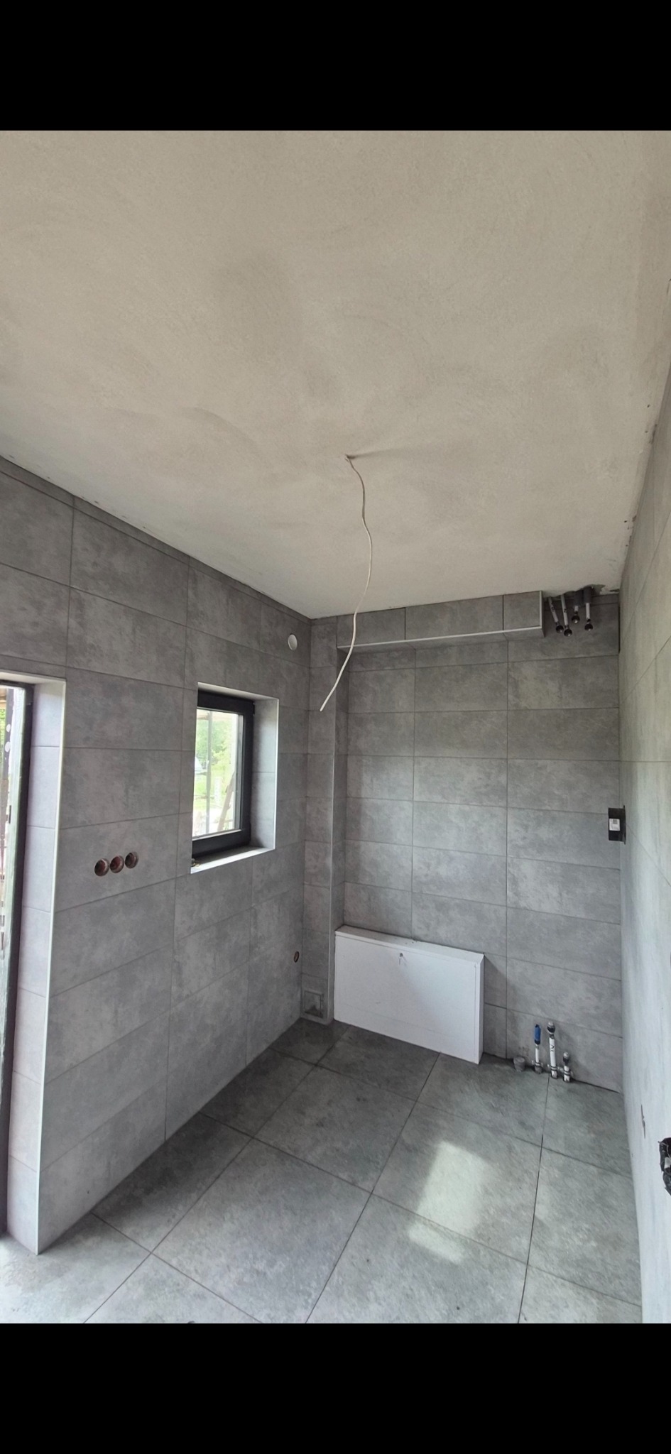 Szara łazienka po remoncie z płytkami na ścianach i podłodze, widoczne okno i grzejnik. Na suficie wystający kabel. Ujęcie z perspektywy narożnika.