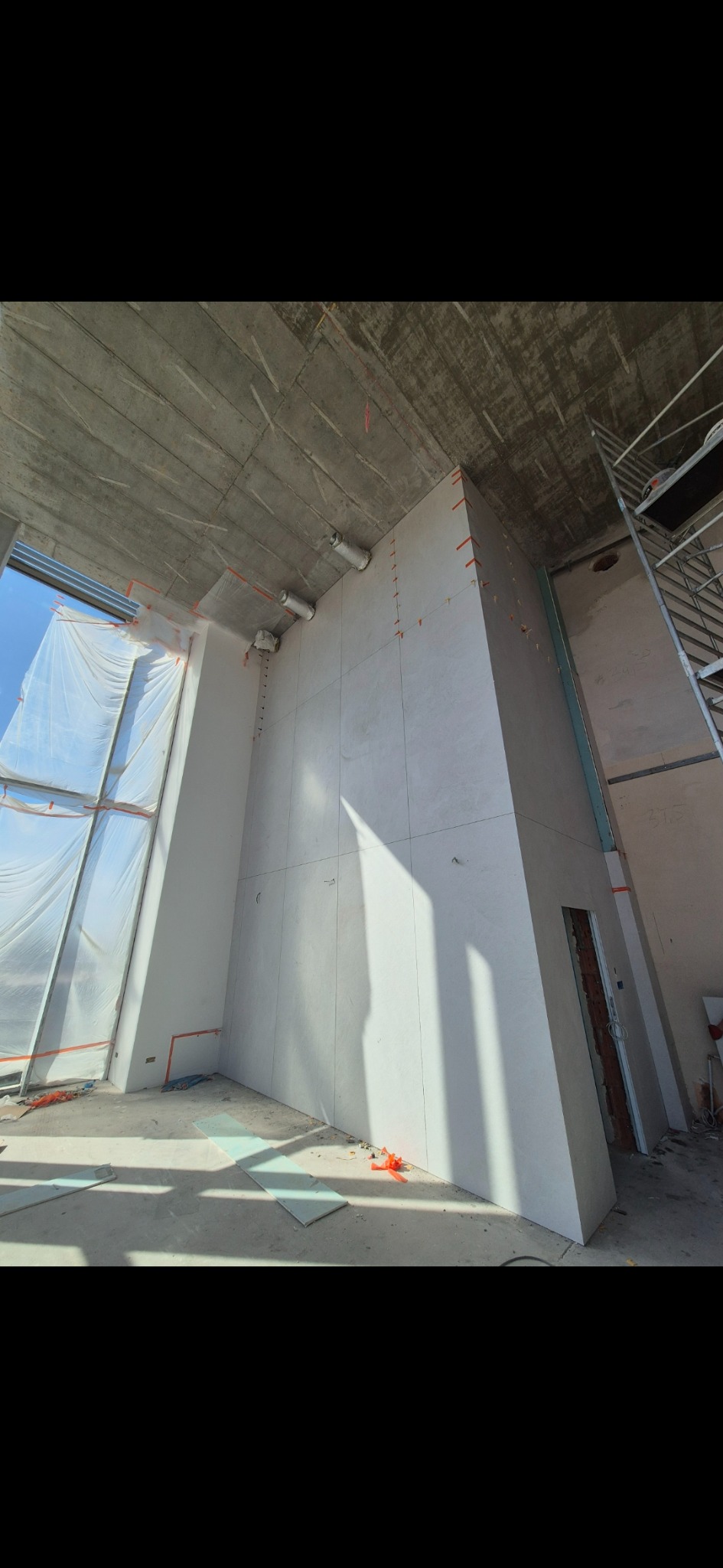 Wnętrze w trakcie prac: ściany wyłożone płytami, widoczne mocowania, surowy betonowy strop, okno z folią ochronną, drabina. Ujęcie z dołu, podkreślające wysokość pomieszczenia.