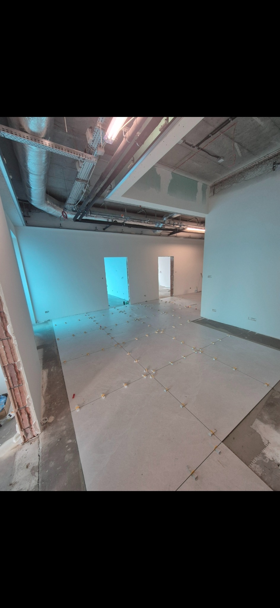 Układanie jasnych płytek podłogowych w przestronnym wnętrzu z widocznymi elementami instalacji pod sufitem i otworami drzwiowymi. Widoczne krzyżyki dystansowe.