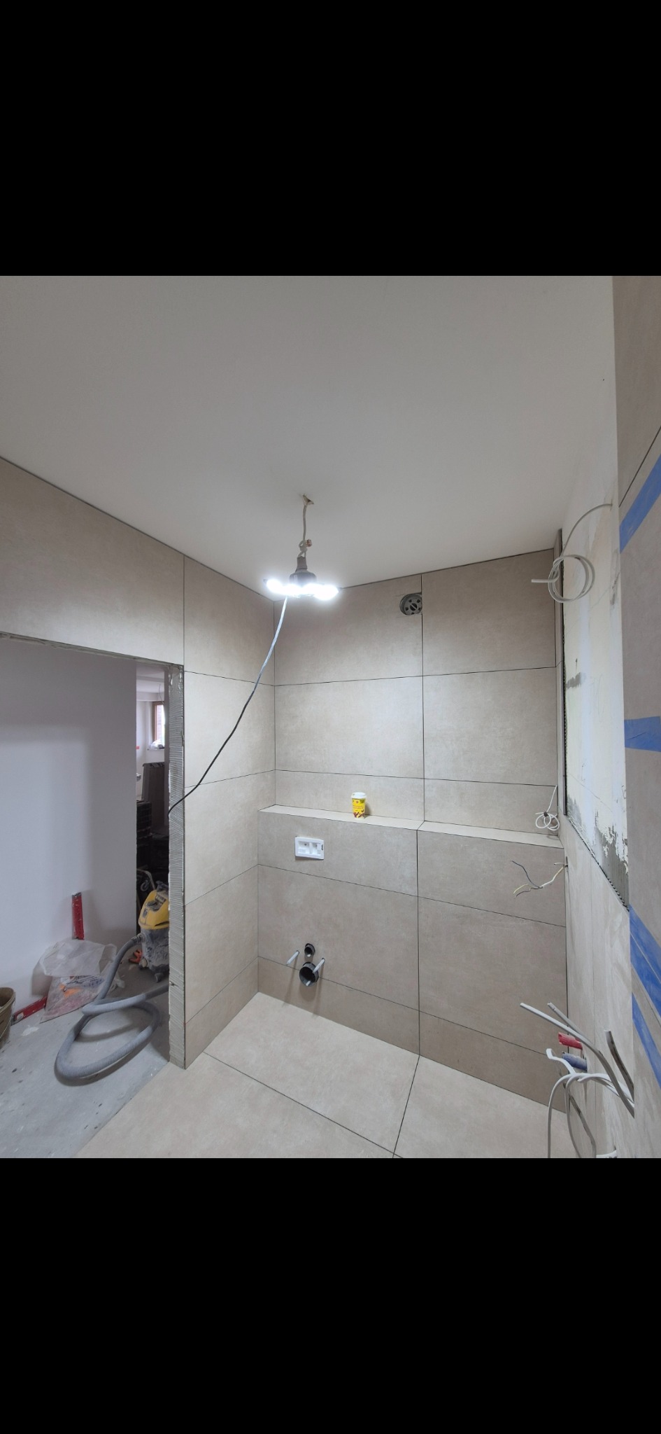 Wnętrze łazienki w trakcie prac wykończeniowych: beżowe płytki na ścianach, widoczne instalacje, oświetlenie robocze i narzędzia. Częściowo wykończone pomieszczenie.