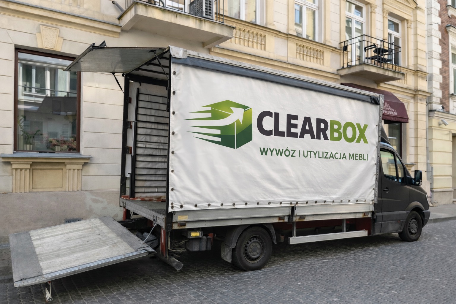 Ciężarówka z otwartą rampą i plandeką z logo firmy Clearbox, oferującej wywóz i utylizację mebli, zaparkowana na tle kamienicy.