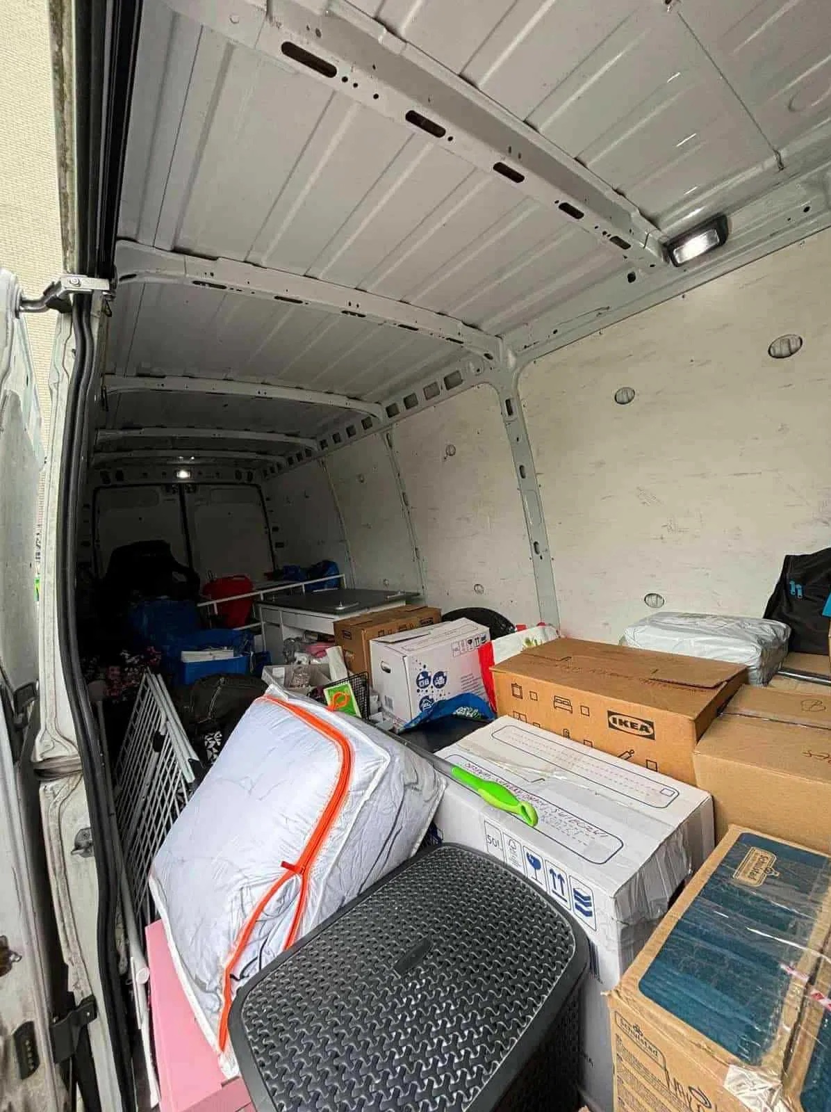 Wnętrze białego busa wypełnione kartonami i różnymi przedmiotami, gotowe do transportu. Widoczne pudełka IKEA i inne pakunki.