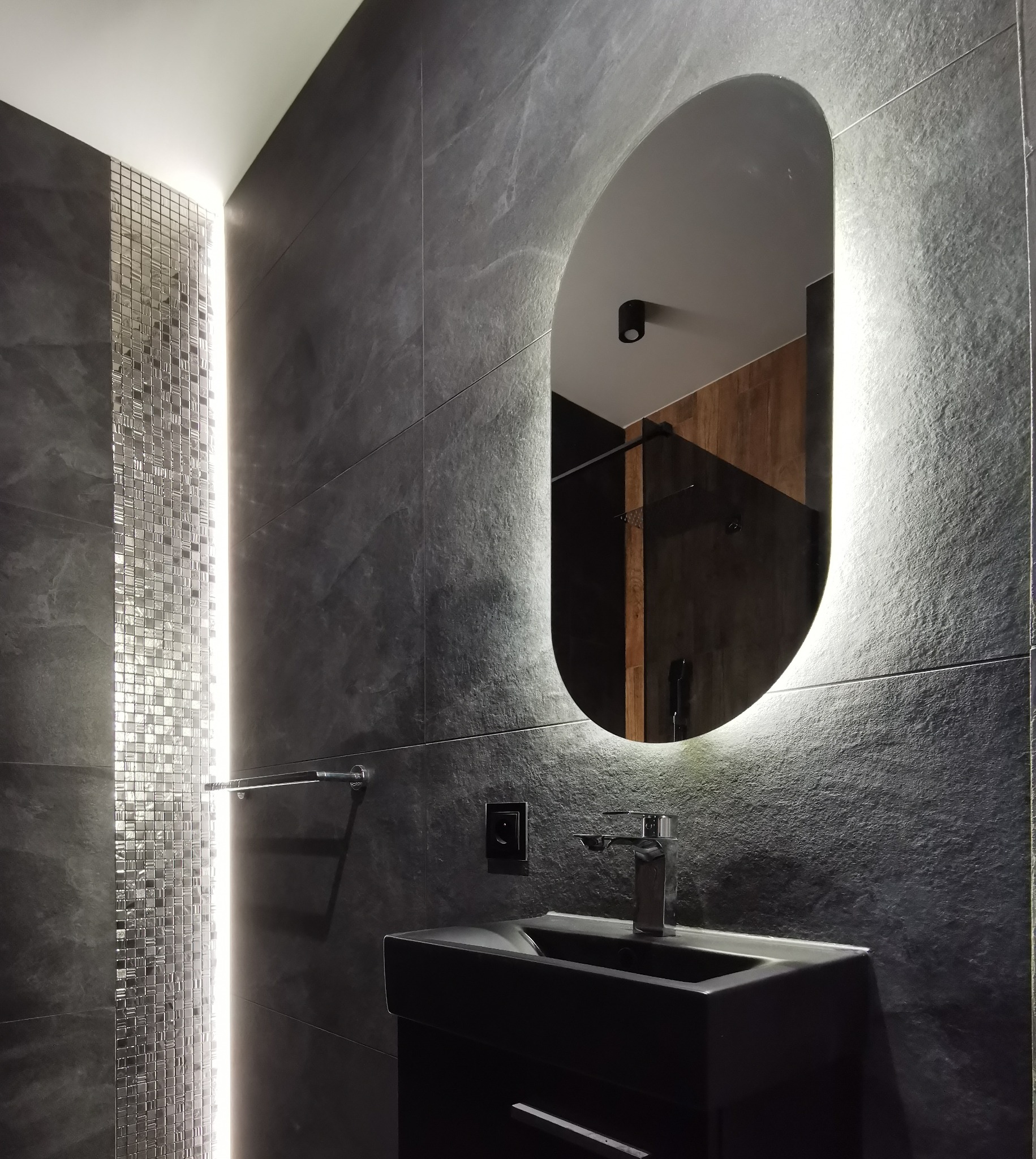 Nowoczesna łazienka z czarnymi płytkami imitującymi kamień, podświetlanym lustrem owalnym i mozaiką z ledowym oświetleniem. Minimalistyczna umywalka nablatowa.