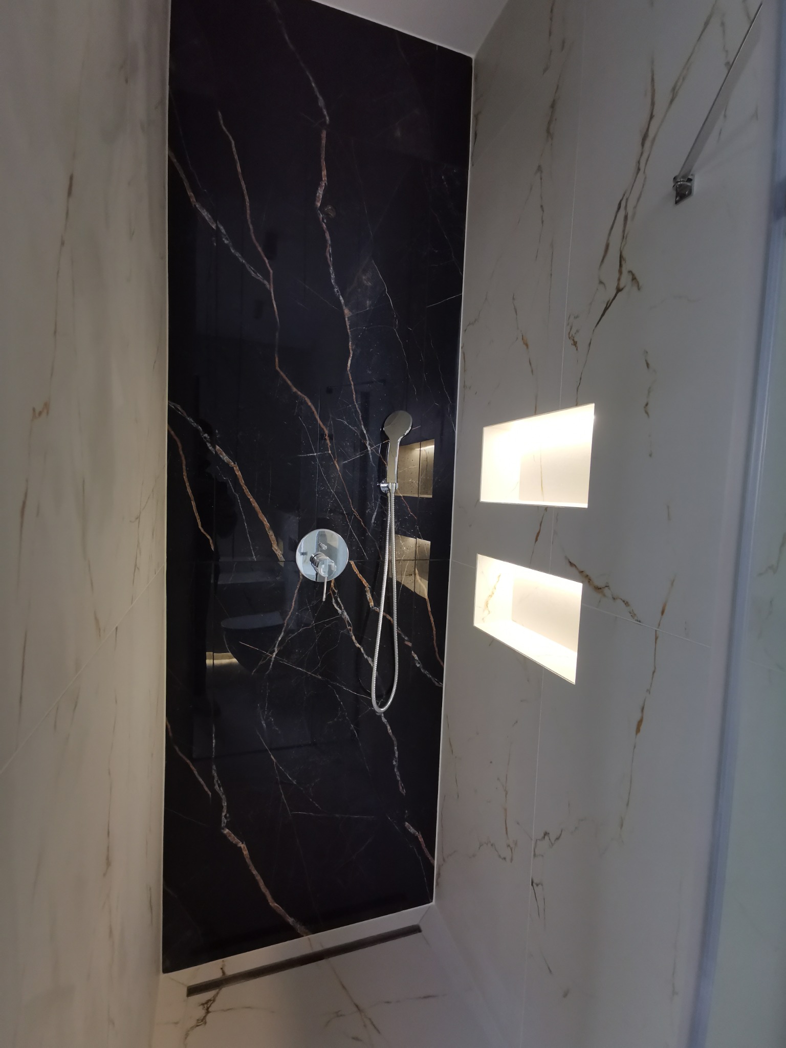 Nowoczesna kabina prysznicowa z czarną, marmurkową ścianą i wnękami z podświetleniem, wykończona płytkami imitującymi biały marmur z delikatnym żyłowaniem.