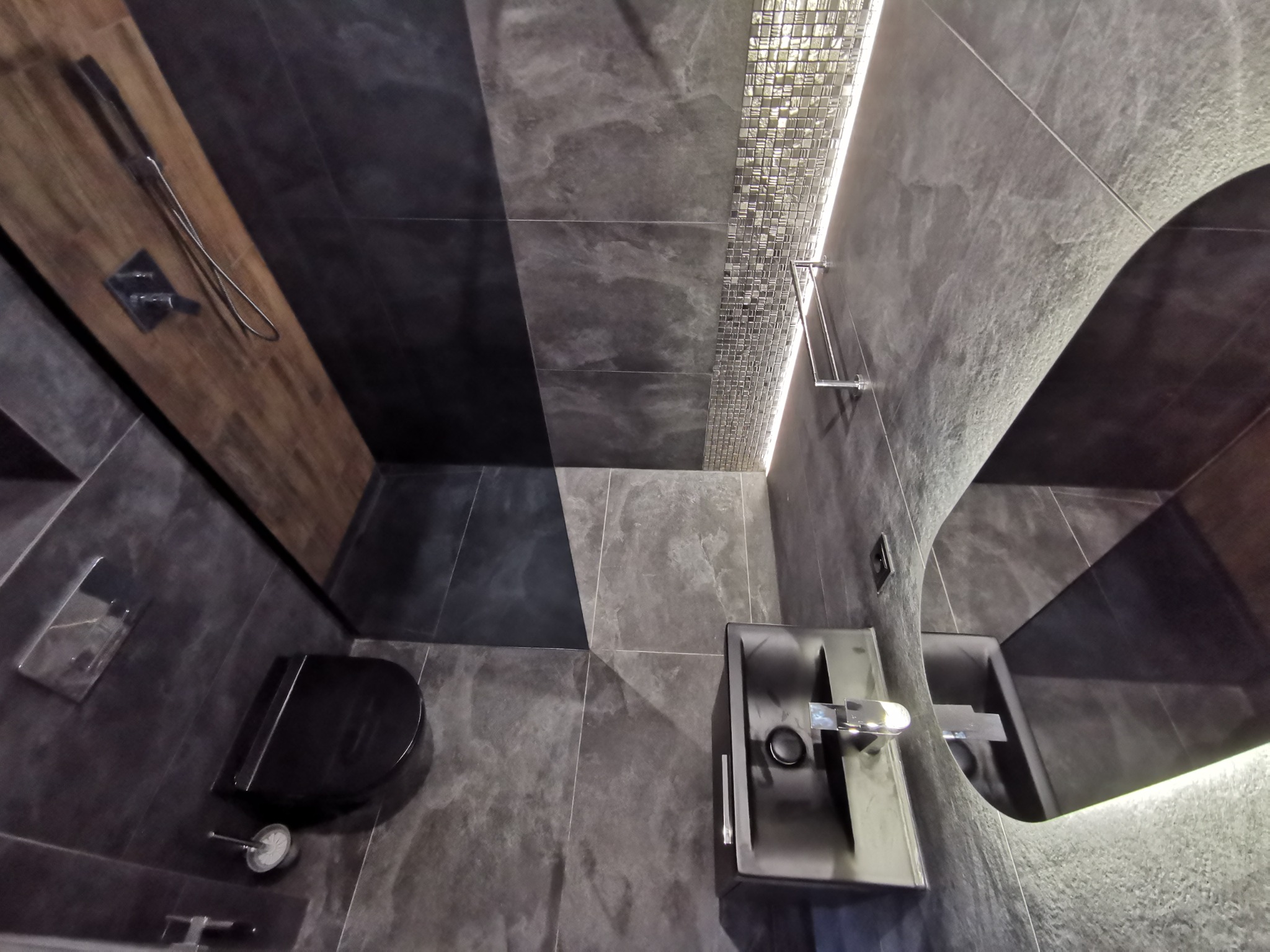 Nowoczesna łazienka z czarną armaturą, szarymi płytkami imitującymi kamień i mozaiką. Umywalka z podświetlanym lustrem. Nietypowy kąt ujęcia.