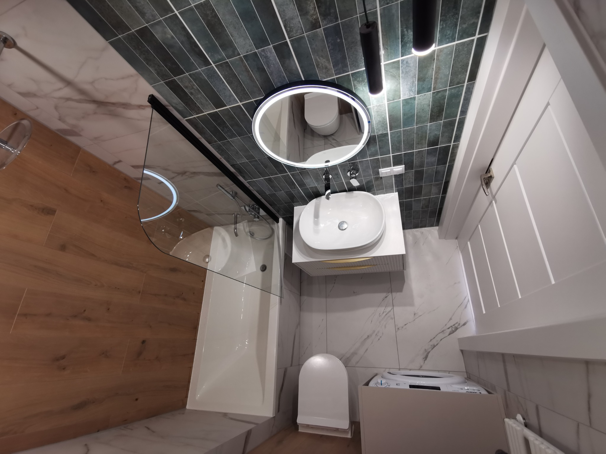 Nowoczesna łazienka z kabiną prysznicową, umywalką z szafką, toaletą i pralką. Ciemne płytki na ścianie kontrastują z jasnymi marmurowymi płytkami i drewnianą podłogą.