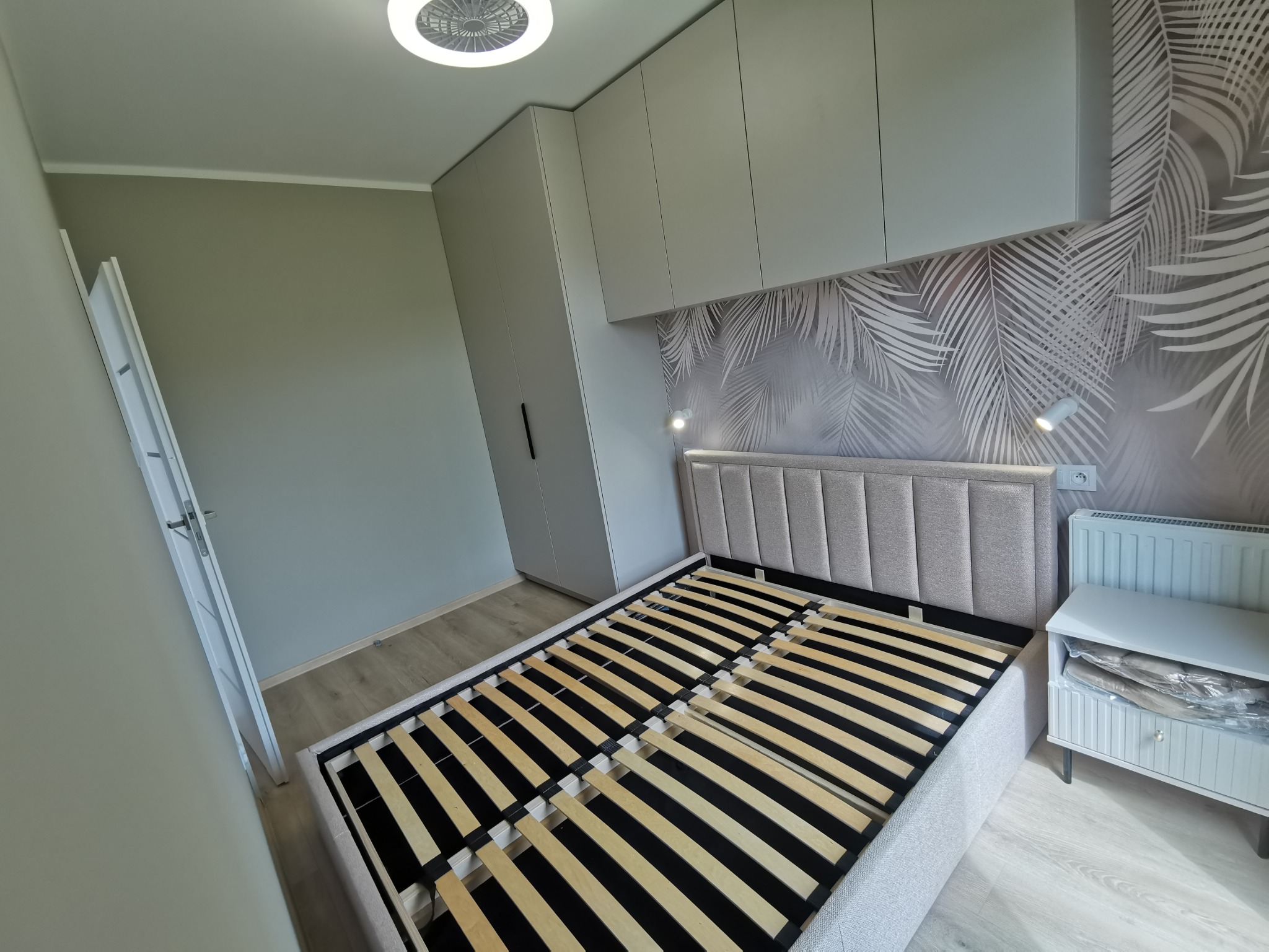 Nowoczesna sypialnia w szarościach: łóżko z tapicerowanym zagłówkiem, szafa w zabudowie, tapeta w liście palmowe i designerskie oświetlenie.