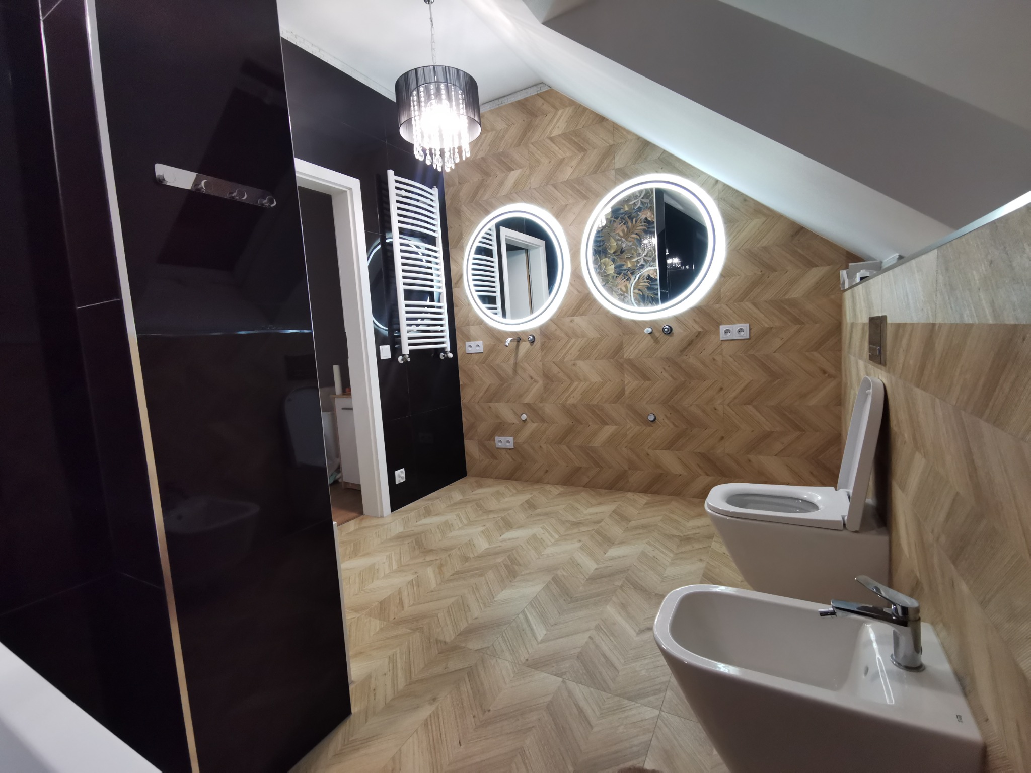 Nowoczesna łazienka na poddaszu z drewnopodobną okładziną, okrągłymi lustrami z podświetleniem LED i czarnymi akcentami. Widoczny bidet i toaleta.