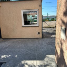 USŁUGI OGÓLNOBUDOWLANE Dawid Nyczka - Szara kostka brukowa ułożona przed budynkiem z oknem, przez które widać osobę. W tle ogrodzenie z siatki i zielony tramwaj. Cień drzewa na kostce.