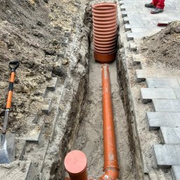USŁUGI OGÓLNOBUDOWLANE Dawid Nyczka - Wykop z ułożonymi rurami kanalizacyjnymi i studzienką, widoczne fragmenty kostki brukowej i łopata. Prace ziemne w trakcie realizacji.