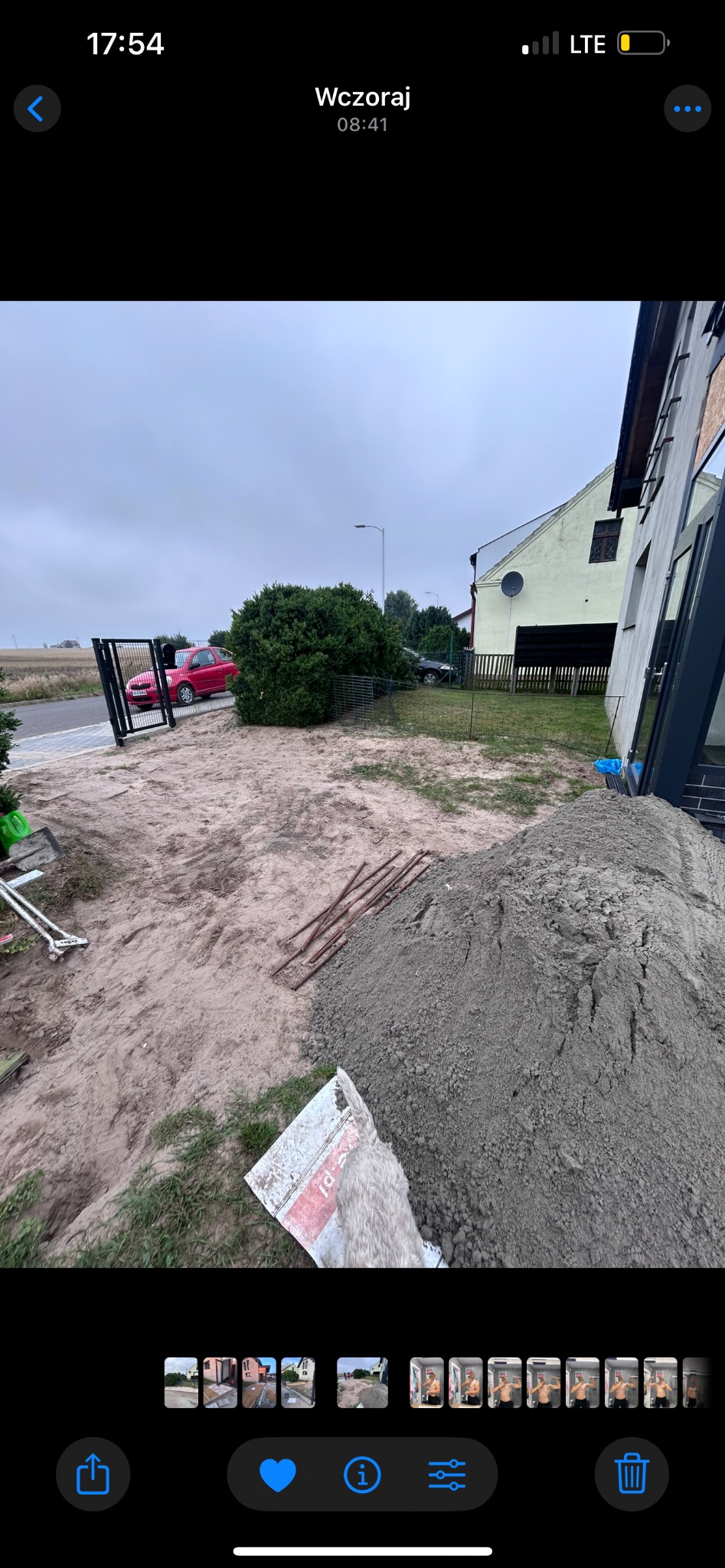 Teren przygotowany pod budowę z widoczną bramą wjazdową, stertą piasku i narzędziami. W tle dom i czerwony samochód. Pochmurne niebo.