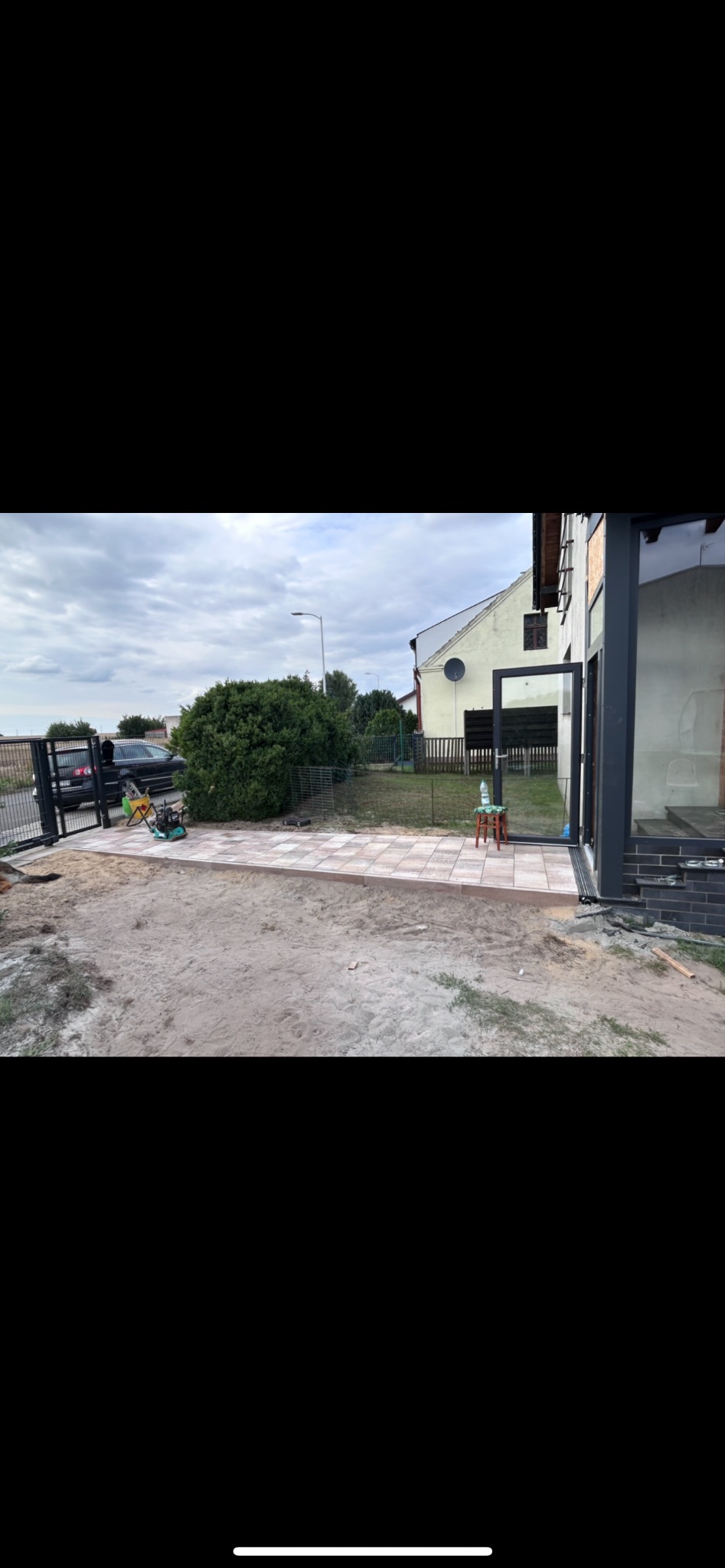 Fragment posesji z częściowo ułożoną kostką brukową w odcieniach beżu i brązu. W tle widoczne ogrodzenie posesji i zieleń. Prace w toku, teren przygotowany pod dalsze układanie.