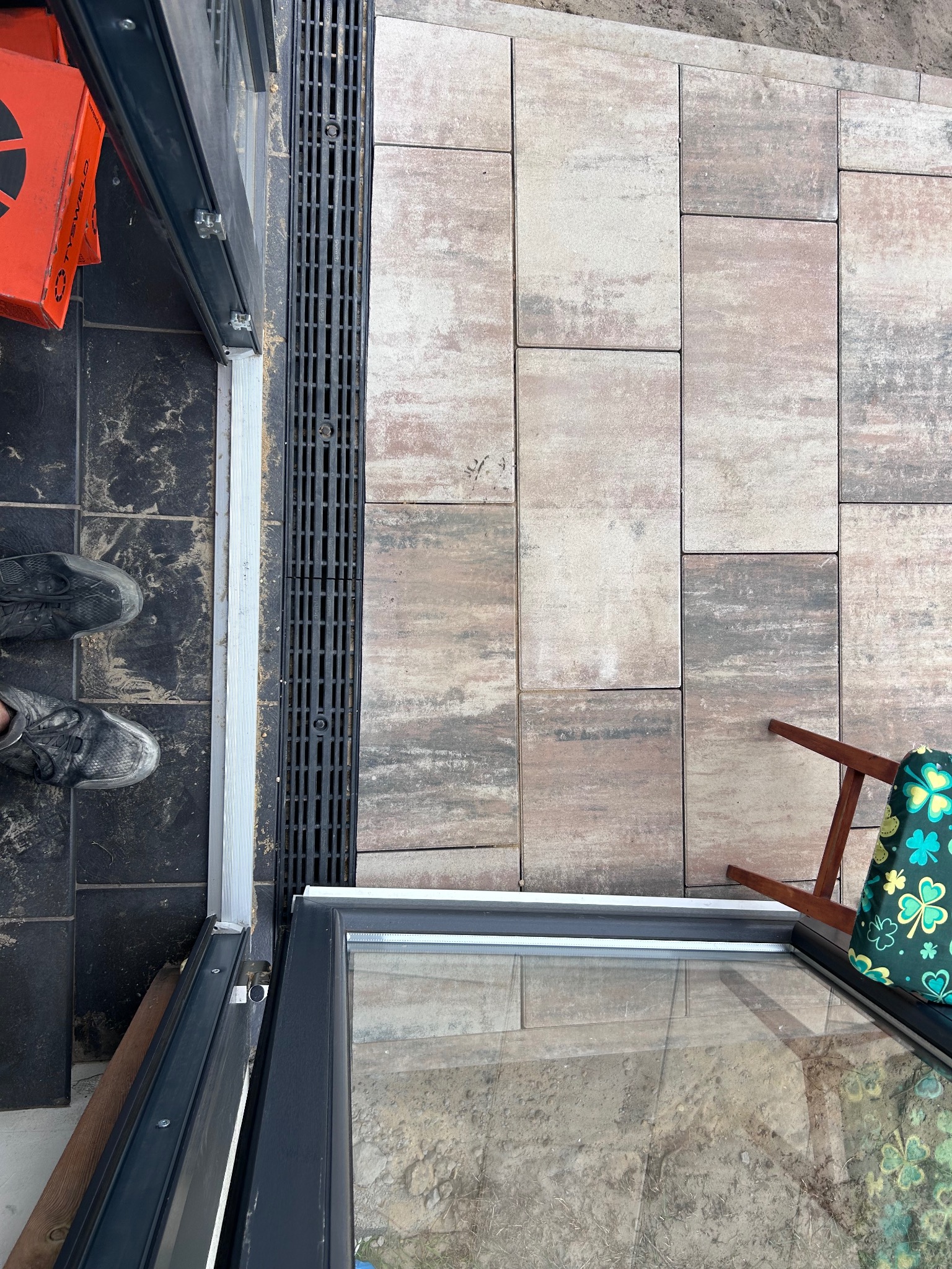 Ułożona kostka brukowa w odcieniach beżu i szarości, oddzielona od ciemnej podłogi odpływem liniowym. Widoczne fragmenty butów i krzesła.