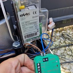 DanEletrick - Ręka technika podłączająca zielony moduł sterujący do napędu bramy FAAC 741 C/ENCODER, widoczne okablowanie i złączki WAGO na tle żwirowego podłoża.