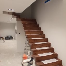 AsBud - Drewniane schody z zabezpieczonymi stopniami w trakcie prac wykończeniowych. Widoczne materiały budowlane i drabina. Oświetlenie sufitowe.