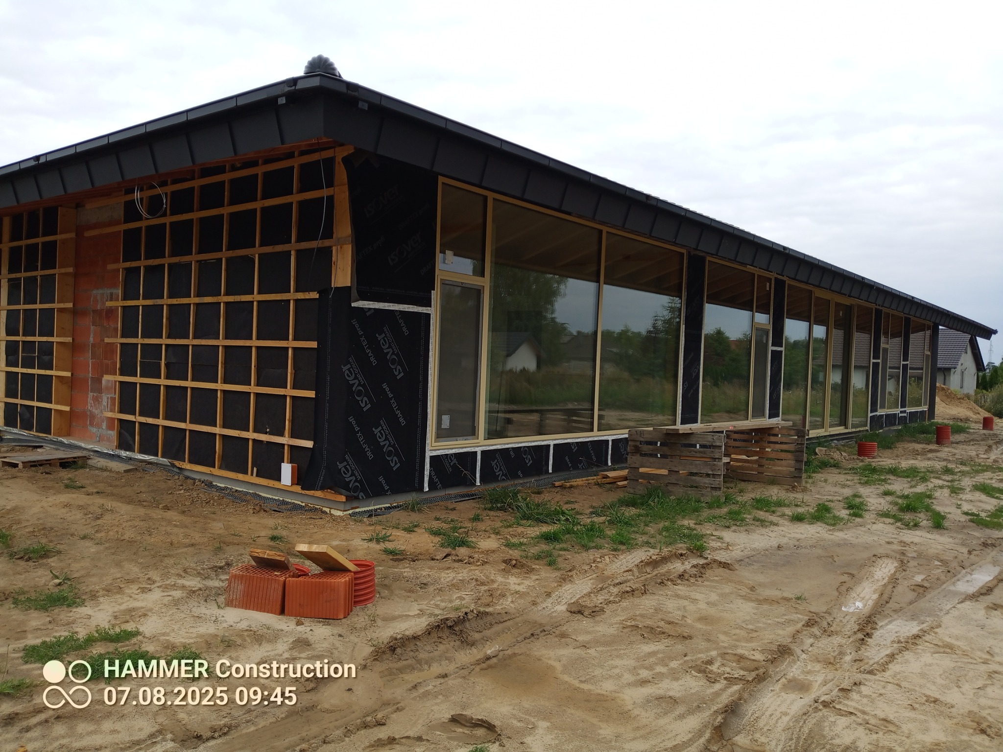 Budynek w trakcie budowy: widoczne okna, drewniana konstrukcja, izolacja. Częściowo otynkowana ściana z cegły. Teren budowy z paletami i materiałami.