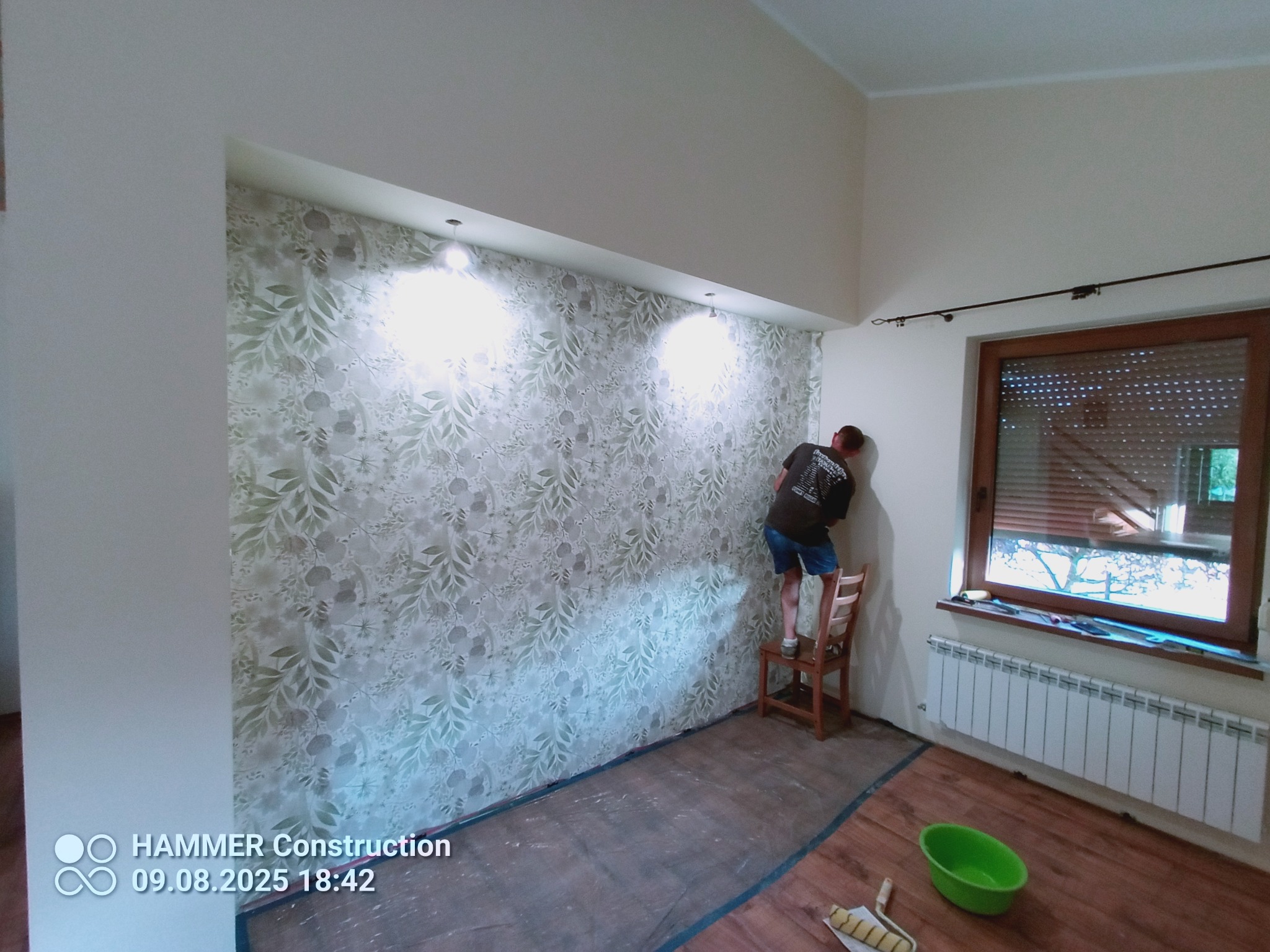 Pracownik na krześle tapetuje ścianę w pokoju z wnęką, widoczne narzędzia malarskie i grzejnik pod oknem. Na podłodze folia ochronna.