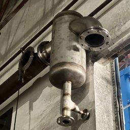 Andrii Vorobiov - Metalowy zbiornik z rurami i kołnierzami, prawdopodobnie element instalacji przemysłowej, zamontowany przy betonowej ścianie. Widoczne ślady spawania.