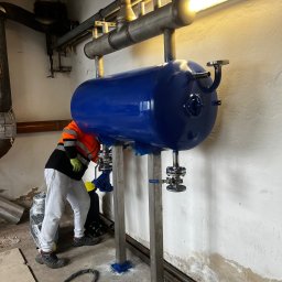 Andrii Vorobiov - Montaż niebieskiego zbiornika ciśnieniowego w kotłowni. Widoczny pracownik w odblaskowej kamizelce podczas instalacji. Rury i elementy instalacji w surowym stanie.