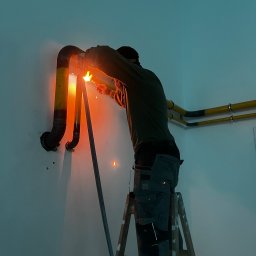 Andrii Vorobiov - Spawanie rur instalacji wewnątrz budynku. Spawacz na drabinie łączy rury, widoczne iskry i żółto-czarne oznaczenia na rurach. Praca w toku, oświetlenie robocze.