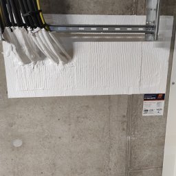 MIWO Jarosław Wojtach - Zabezpieczenie ogniochronne przejść kablowych przez ścianę betonową, z widoczną metalową konstrukcją wsporczą i etykietą informacyjną PYRO-SAFE.