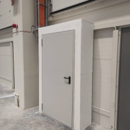 MIWO Jarosław Wojtach - Szara, metalowa, przeciwpożarowa brama w betonowej obudowie, zamontowana w hali przemysłowej. Nad bramą widoczne kable w korytkach. W tle brama garażowa.