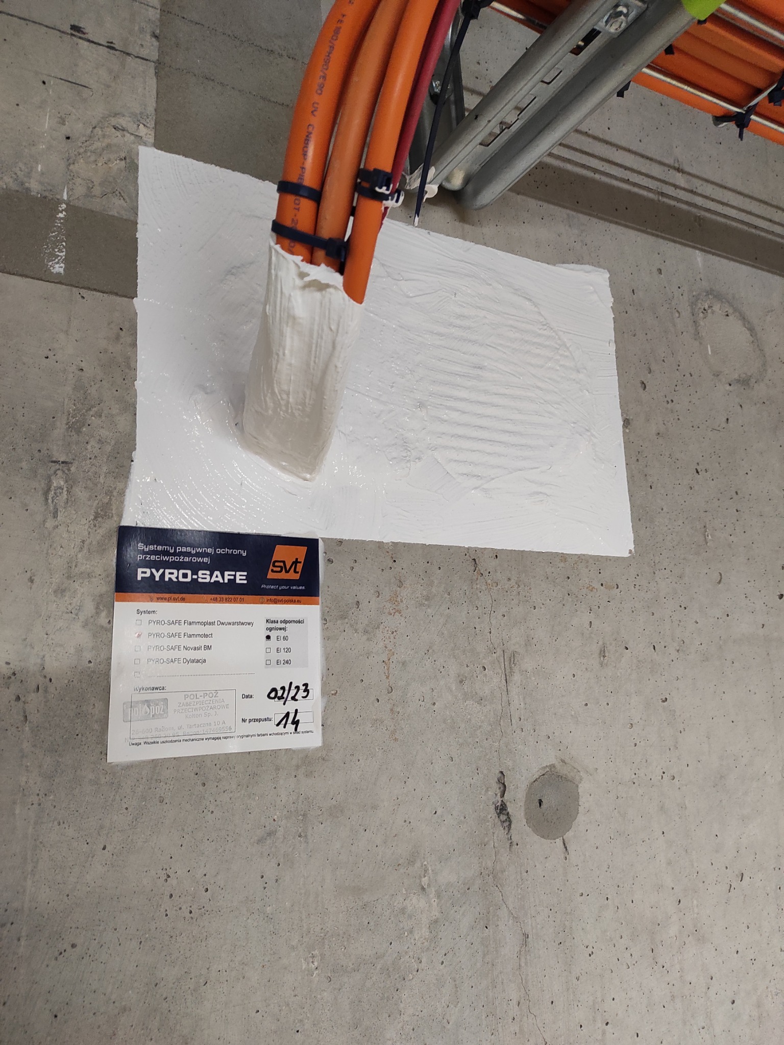 Zabezpieczenie przeciwpożarowe przepustu kablowego w ścianie betonowej, wykonane systemem PYRO-SAFE. Widoczna etykieta z datą wykonania i numerem przepustu.