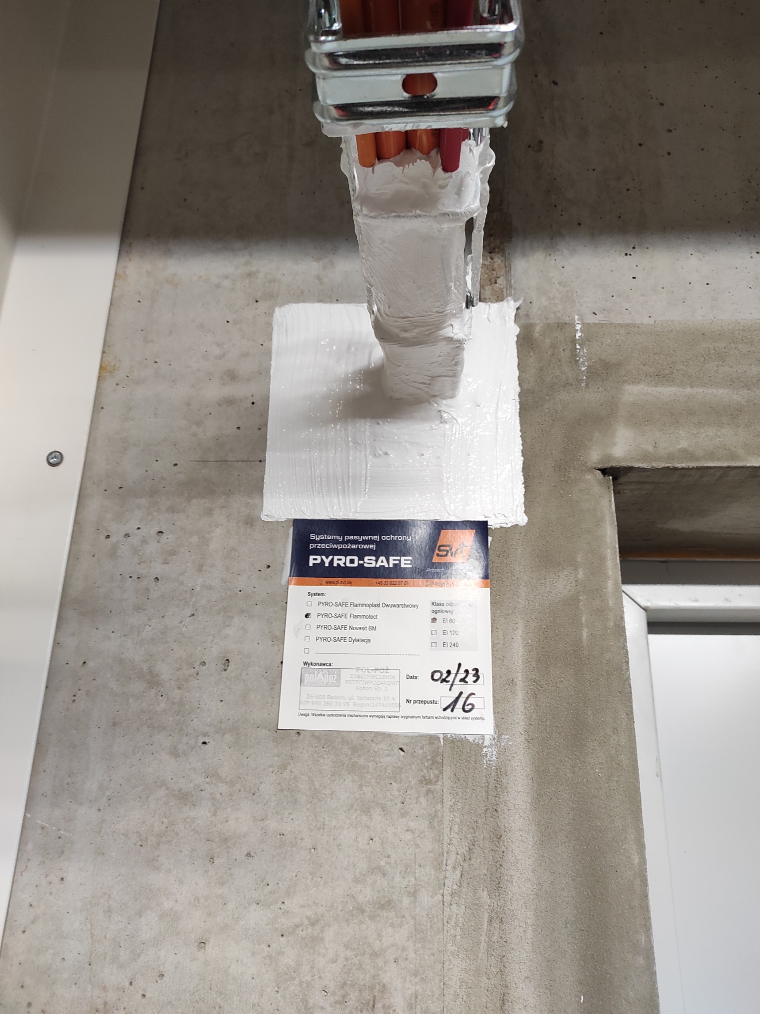 Zabezpieczenie ogniochronne przepustu kablowego w ścianie betonowej, uszczelnione białą masą. Widoczna etykieta systemu PYRO-SAFE z datą wykonania 02/23 i numerem przepustu 16.