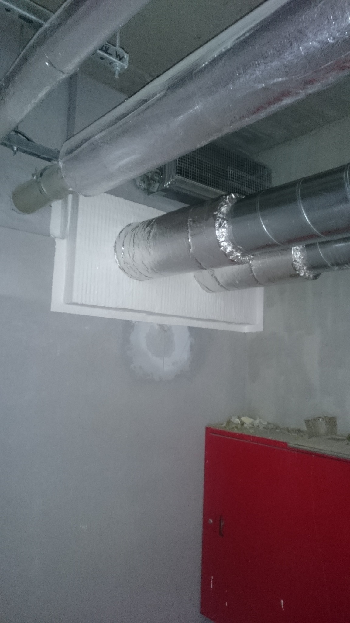 Instalacja wentylacyjna z izolacją termiczną na suficie, widoczna czerwona szafka. Ujęcie z dołu, sufit z elementami instalacji.