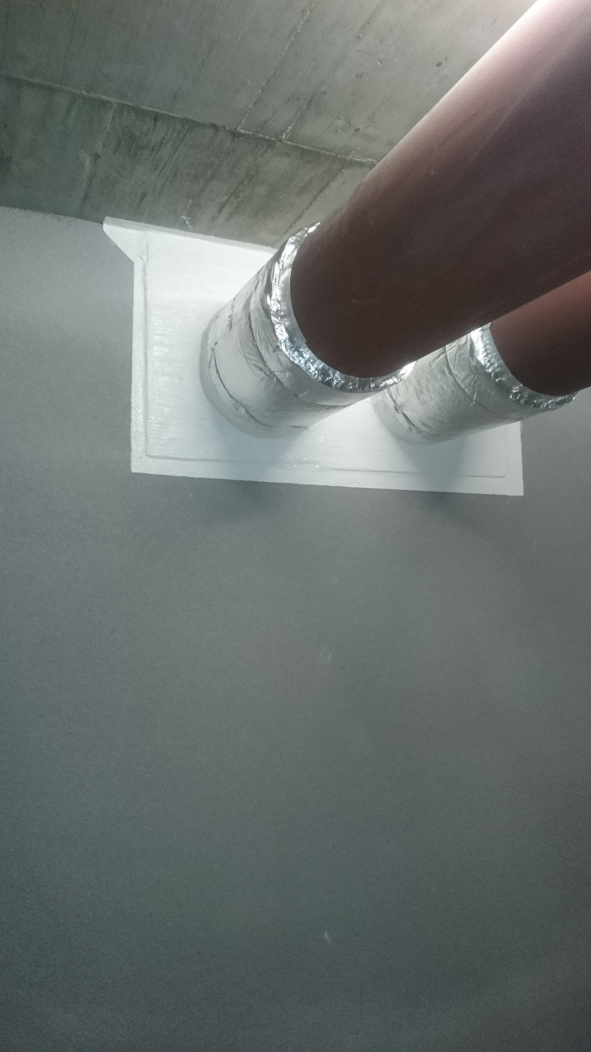 Dwa brązowe przewody, owinięte srebrną folią, przechodzą przez białą płytę przymocowaną do szarej ściany i betonowego sufitu. Widok z dołu.