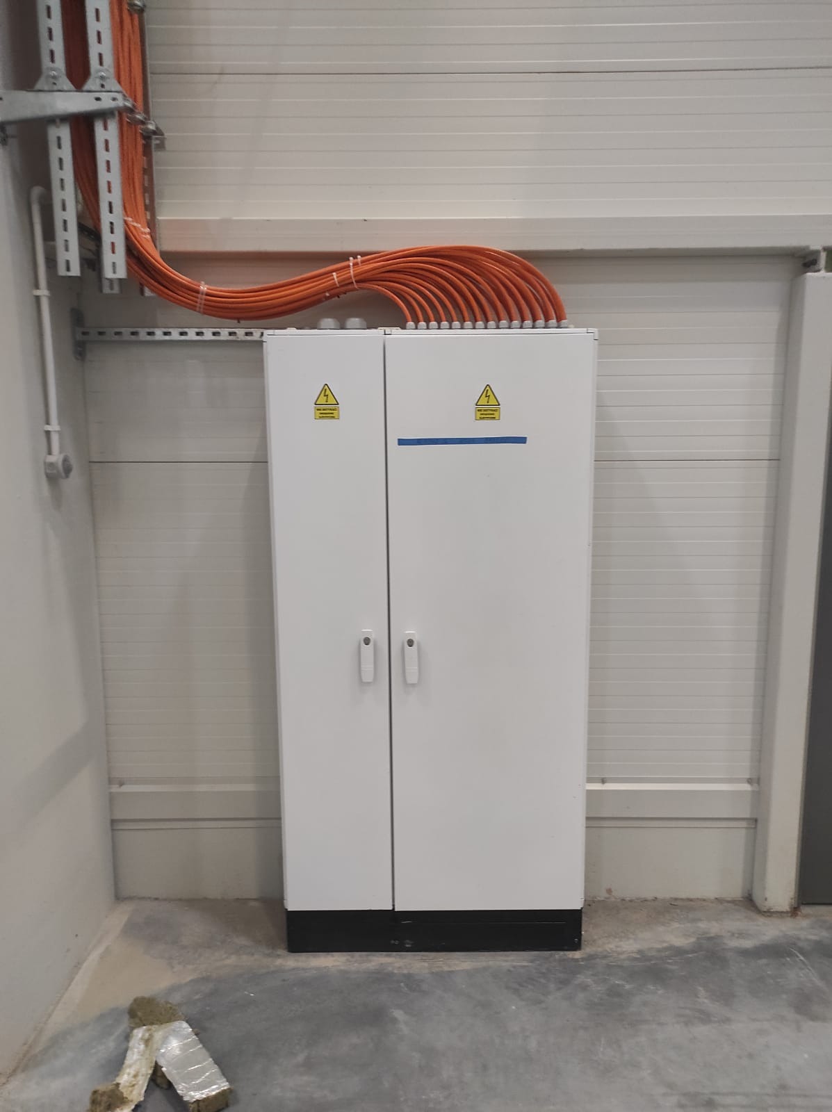 Biała szafa elektryczna z pomarańczowymi kablami na górze, umieszczona w hali z szarymi ścianami i betonową podłogą. Widoczne znaki ostrzegawcze.