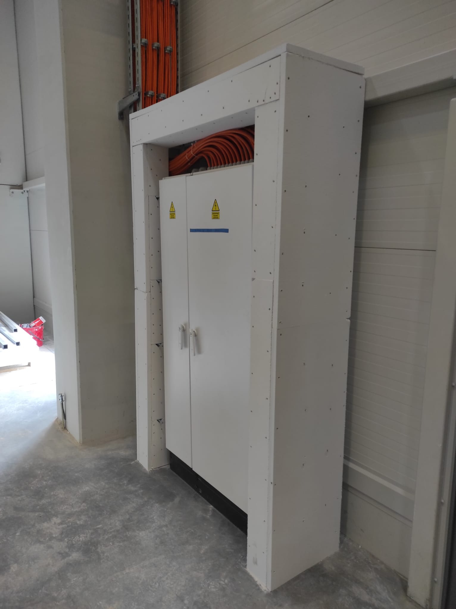 Biała szafa elektryczna z pomarańczowymi kablami w obudowie z płyt gipsowo-kartonowych, widoczne znaki ostrzegawcze. Szare betonowe podłoże, surowy stan wykończenia.